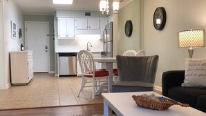Un salon moderne avec un canapé blanc, une table à manger agrémentée d’un panier, et un coin cuisine doté de meubles blancs et d’un réfrigérateur.