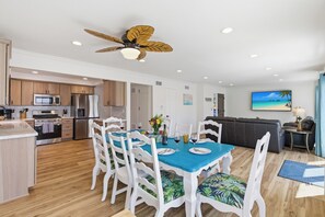 Una cocina moderna con microondas, horno y refrigerador, un comedor con una mesa azul y sillas blancas, y una sala de estar con sofá y televisor.
