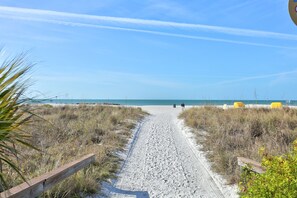 A sandy path leading to a beach with a wooden fence on one side and yellow chairs on the other.