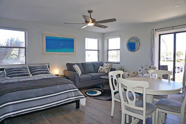 A room with a bed, a sofa, a dining table with chairs, a ceiling fan, and large windows. - 🌺 Upgraded Studio with Garden-View Balcony — Gulf Winds Resort (Saint Pete Beach)
