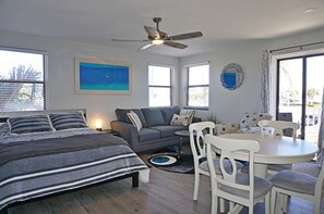 Quarto com cama, sofá, mesa de jantar com cadeiras, ventilador de teto e janelas amplas.