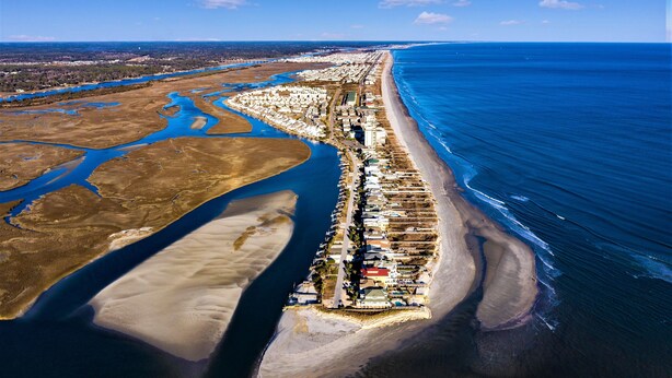 Vue aérienne d’un complexe immobilier situé sur la côte, avec une longue plage, des étendues d’eau adjacentes et un ensemble dense de bâtiments.