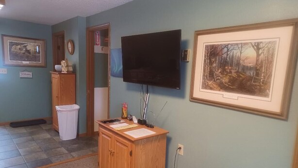 A room with a wooden desk, a flat-screen TV mounted on the wall, a framed landscape painting, and a door leading to another room.