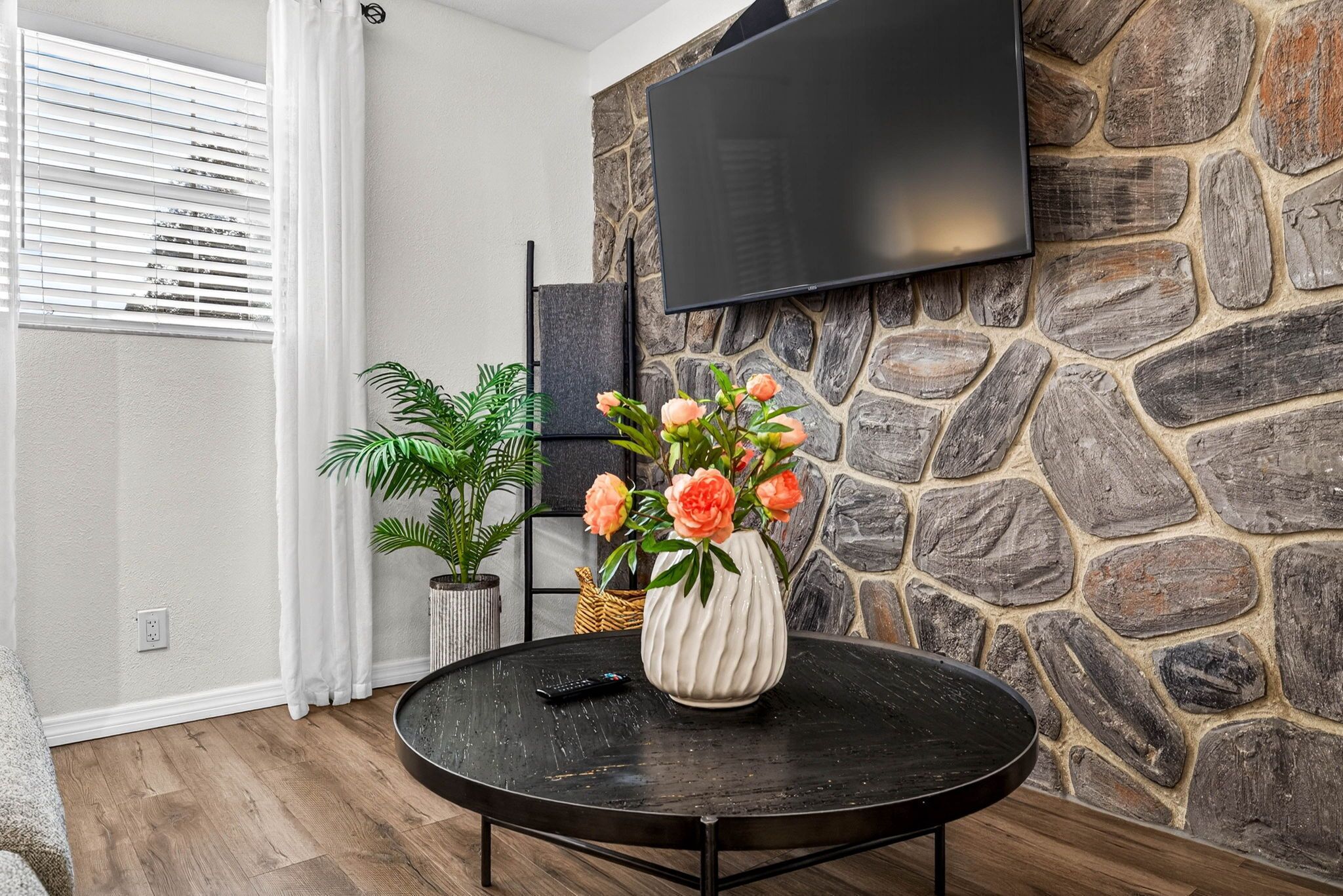 En stue med en stenvæg, et ophængt fjernsyn, et rundt sort bord med en vase blomster og en potteplante.
