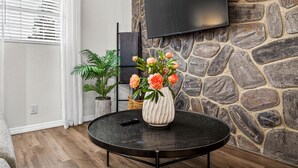 En stue med en stenvæg, et ophængt fjernsyn, et rundt sort bord med en vase blomster og en potteplante.