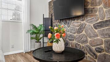 En stue med en stenvæg, et ophængt fjernsyn, et rundt sort bord med en vase blomster og en potteplante.