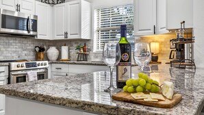 Une cuisine avec des meubles blancs, un plan de travail en granit, une bouteille de vin, un verre et une planche à découper avec du fromage et des raisins.