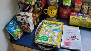 Una encimera de cocina con diversos alimentos, una cafetera, una taza y un juego de búsqueda de palabras.