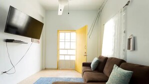 Un salón con un televisor de pantalla plana montado en la pared, un sofá marrón con cojines, una alfombra azul estampada y una puerta que da al exterior.