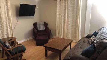 Een woonkamer met houten vloer, een bruine bank, een houten salontafel, een schommelstoel, een fauteuil en een plafondventilator.