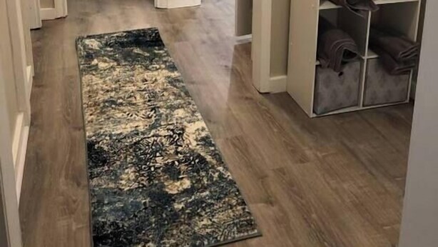Ein Flur mit Holzboden, einem gemusterten Teppich, einem Bett mit Kopfteil, einem Schreibtisch mit Lampe und einem Regal mit gefalteten Decken.
