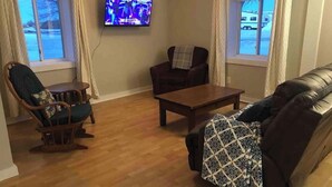 Ein Wohnzimmer mit Holzboden, einem Ledersofa, einem Holzsesseln, einer karierten Decke auf dem Sessel, einem Couchtisch und einem Fernseher an der Wand.