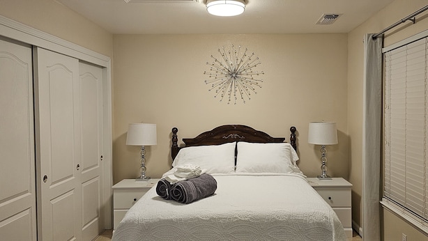 A neatly made bed with white linens, two bedside lamps, a decorative wall piece, and a window with blinds.