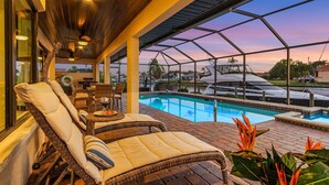 A covered outdoor area with a pool, lounge chairs, and a view of a boat and palm trees. - Amazing water views, heated pool & spa, Tiki & kayaks -Villa SeaOarNo - Roelens Vacations (Cape Coral)
