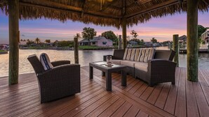 A wooden deck with wicker furniture and a thatched roof, overlooking a body of water and a house. - Amazing water views, heated pool & spa, Tiki & kayaks -Villa SeaOarNo - Roelens Vacations (Cape Coral)