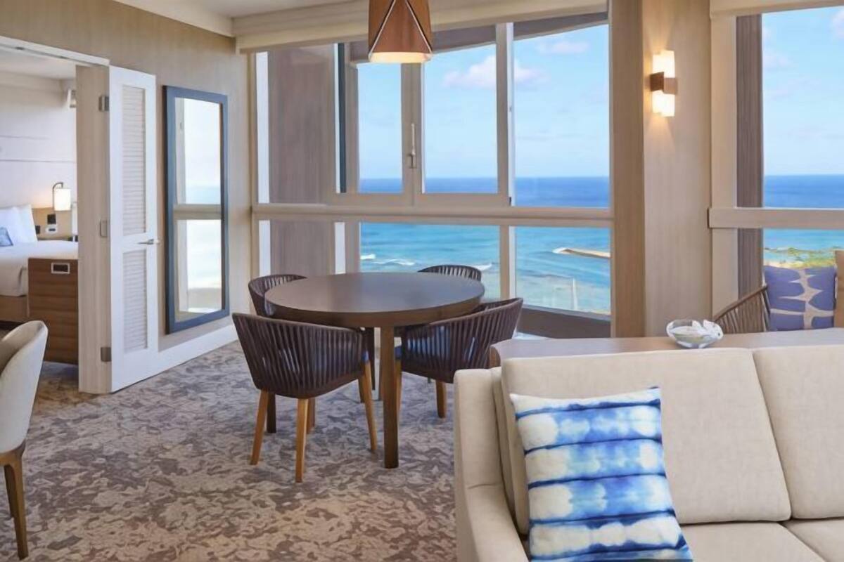 A room with a large window, a round table, and a sofa with a patterned pillow.