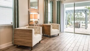 Une pièce spacieuse avec un plancher en bois, deux fauteuils, une table d’appoint avec une lampe et une grande fenêtre avec des rideaux.