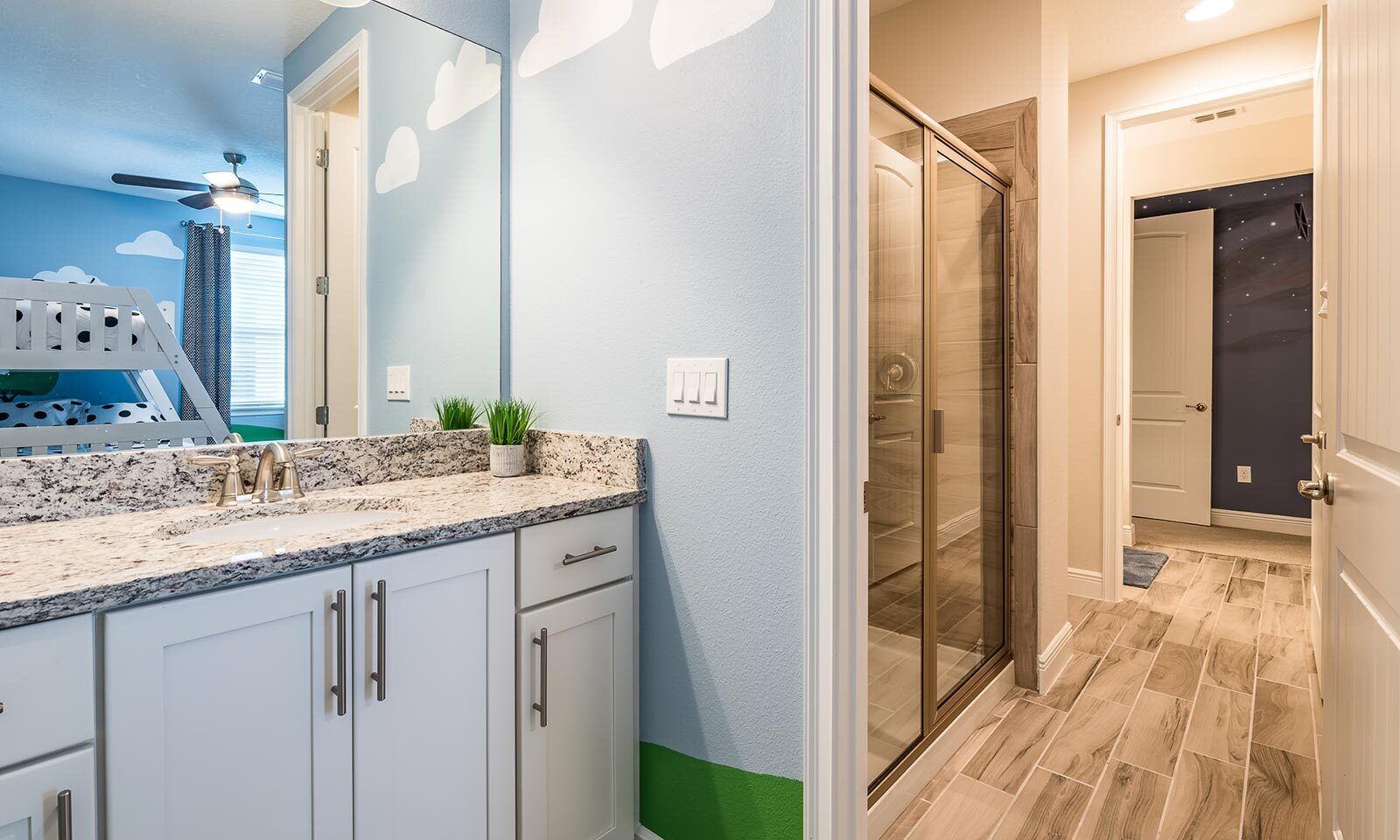 Une salle de bain avec une porte de douche en verre, un lavabo blanc doté d’un plan de travail en granit et un ventilateur de plafond.