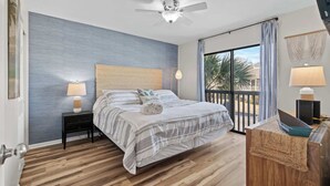 Une chambre à coucher comprenant un lit, une table de chevet, un ventilateur de plafond, une fenêtre avec des rideaux et une vue sur des palmiers.