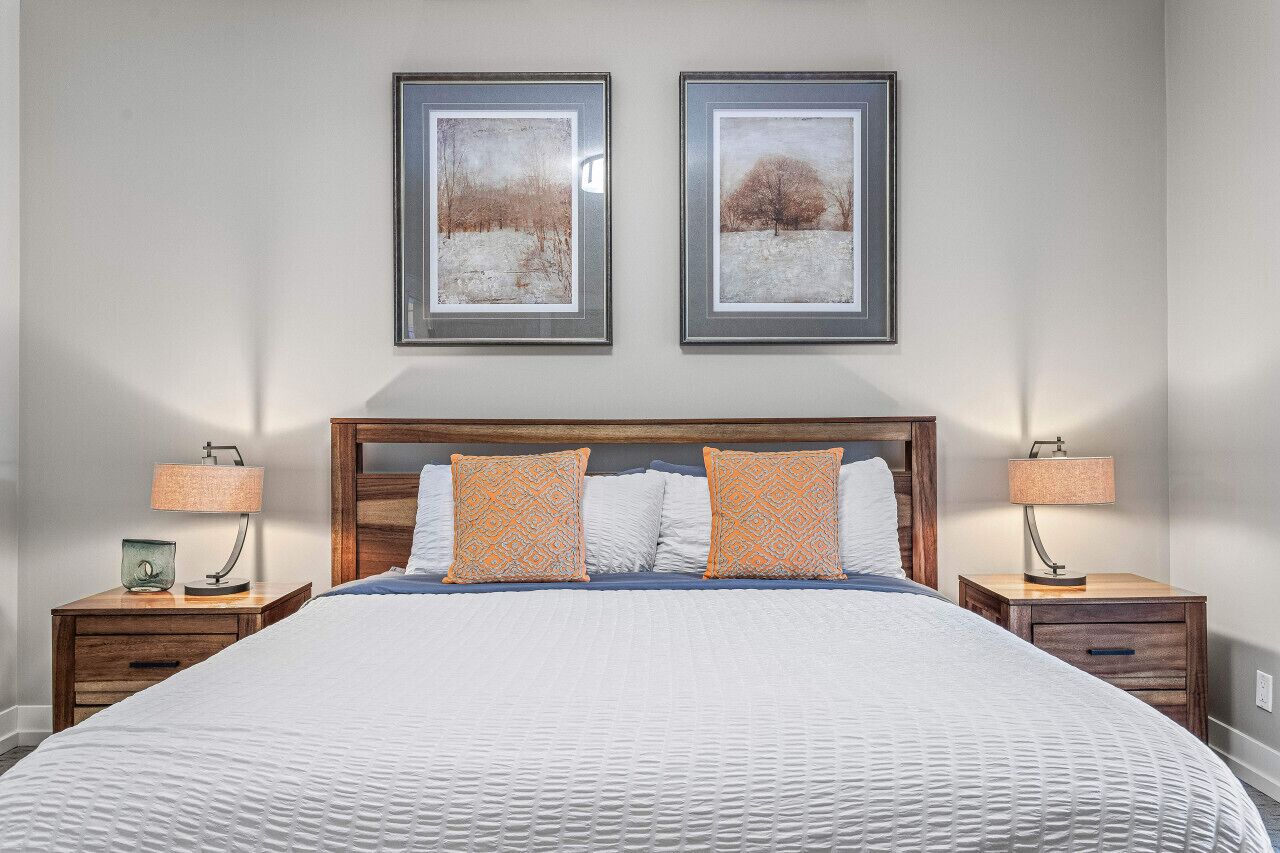 Una cama bien hecha con colcha blanca, dos almohadas a juego con estampado, y un cabecero de madera. Encima de la cama, en la pared, hay dos cuadros enmarcados, y dos mesitas de noche con lámparas y pequeños objetos decorativos.