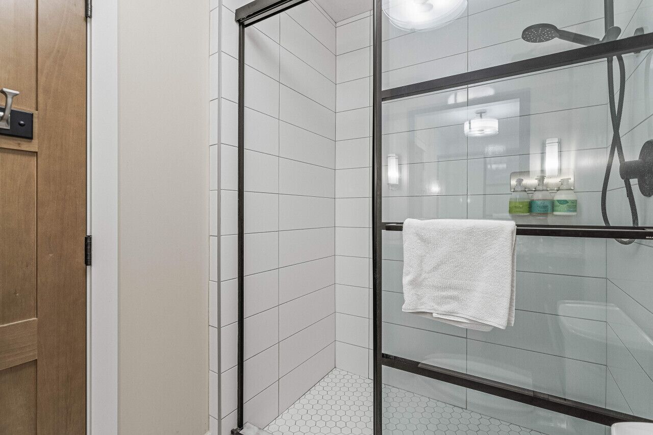 Un baño moderno con cabina de ducha de vidrio, paredes de azulejos blancos y una puerta de madera.