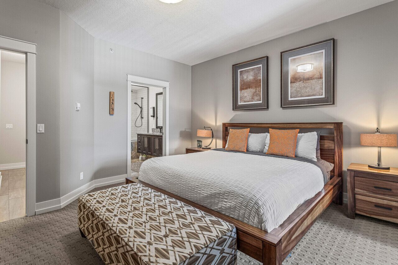 Une chambre avec un grand lit, une tête de lit en bois, deux tables de chevet avec des lampes, un banc à motifs et une porte donnant sur une salle de bain.