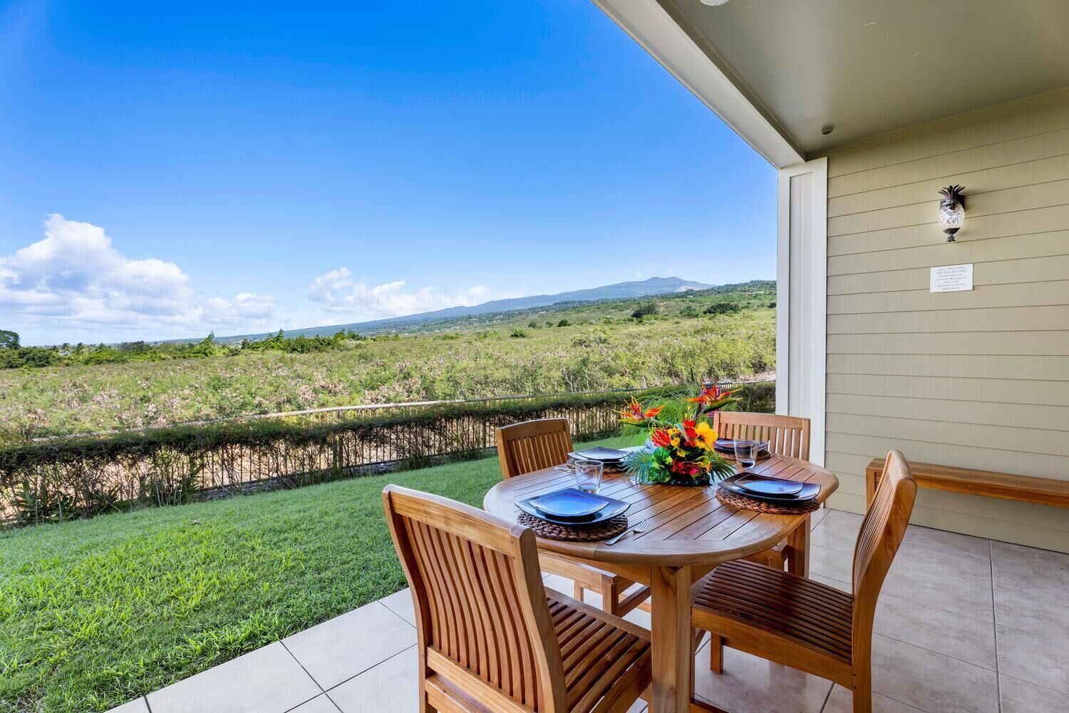 A wooden table set for dining with chairs, a view of a grassy field, and a distant landscape.