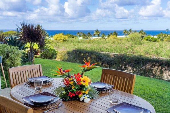 Une table ronde en bois, dressée pour quatre personnes, avec des assiettes, des couverts et un centre de table composé de fleurs tropicales.