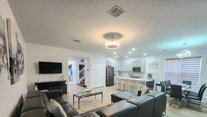 A modern living room with a sofa, a glass coffee table, and a TV. There is a kitchen area with a dining table and chairs. - BELLA VIDA (891MB) - AMAZING 5BR 4BA HOME*POOL&SPA*GATED RESORT (Kissimmee)