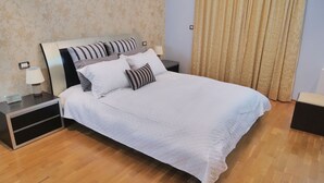 A neatly made bed with white bedding and striped pillows, a bedside table with a lamp, and a wooden floor. - Villa Omega Kefalonia (Karavados)