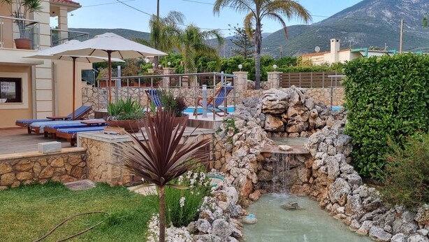 A pool area with lounge chairs, a small waterfall, and surrounding greenery. - Villa Omega Kefalonia (Karavados)