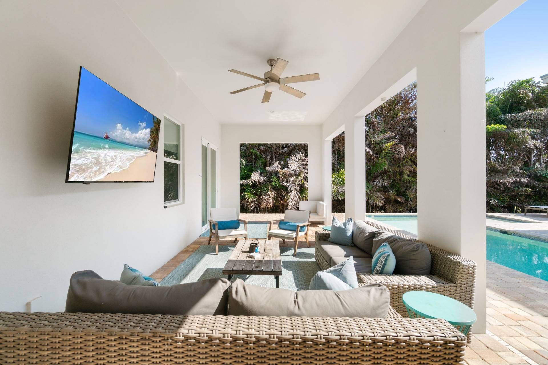 A sunlit indoor space with a large TV, wicker furniture, a ceiling fan, and a view of a pool and tropical plants.