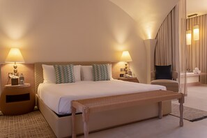 Une chambre d’hôtel avec un grand lit, deux tables de chevet avec des lampes, un banc, une chaise et une grande fenêtre avec des rideaux.