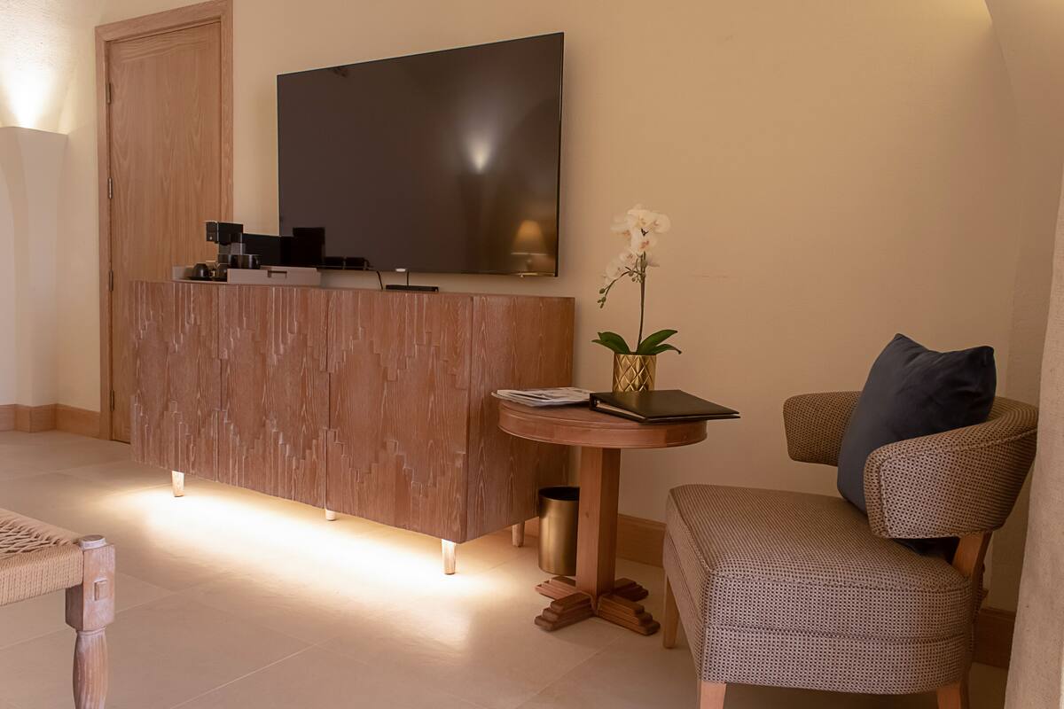 Een moderne woonkamer met een houten kast, een flatscreen televisie, een klein rond tafeltje met een boek en een bloemenvaas, en een gestreept fauteuil.