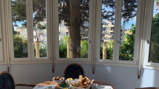 Ein Esstisch mit Frühstücksangeboten, umgeben von Stühlen, in einem Raum mit großen Fenstern mit Blick auf eine Wohngegend.