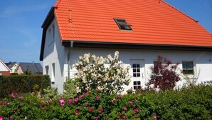 A white house with a red roof, a small window, and a garden with pink flowers.