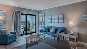 Una sala de estar moderna con una mesa de comedor de vidrio, sofás azules, una lámpara blanca y vistas de la ciudad a través de amplias ventanas.
