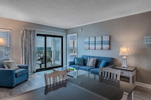 A modern living room with a glass dining table, blue sofas, a white lamp, and a city view through large windows.