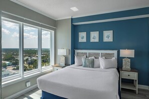 A bedroom with a large window, a bed with white bedding, a nightstand with a lamp, and a blue accent wall. - Coastal Luxe | Palace Twenty-One Thirteen (Myrtle Beach)
