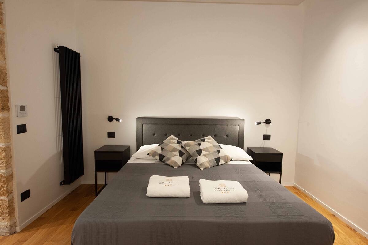 Een net opgemaakt bed met een grijze sprei, twee kussens en twee gevouwen handdoeken. Twee nachtkastjes met lampen, een radiator aan de muur en een bakstenen muur aan de zijkant.