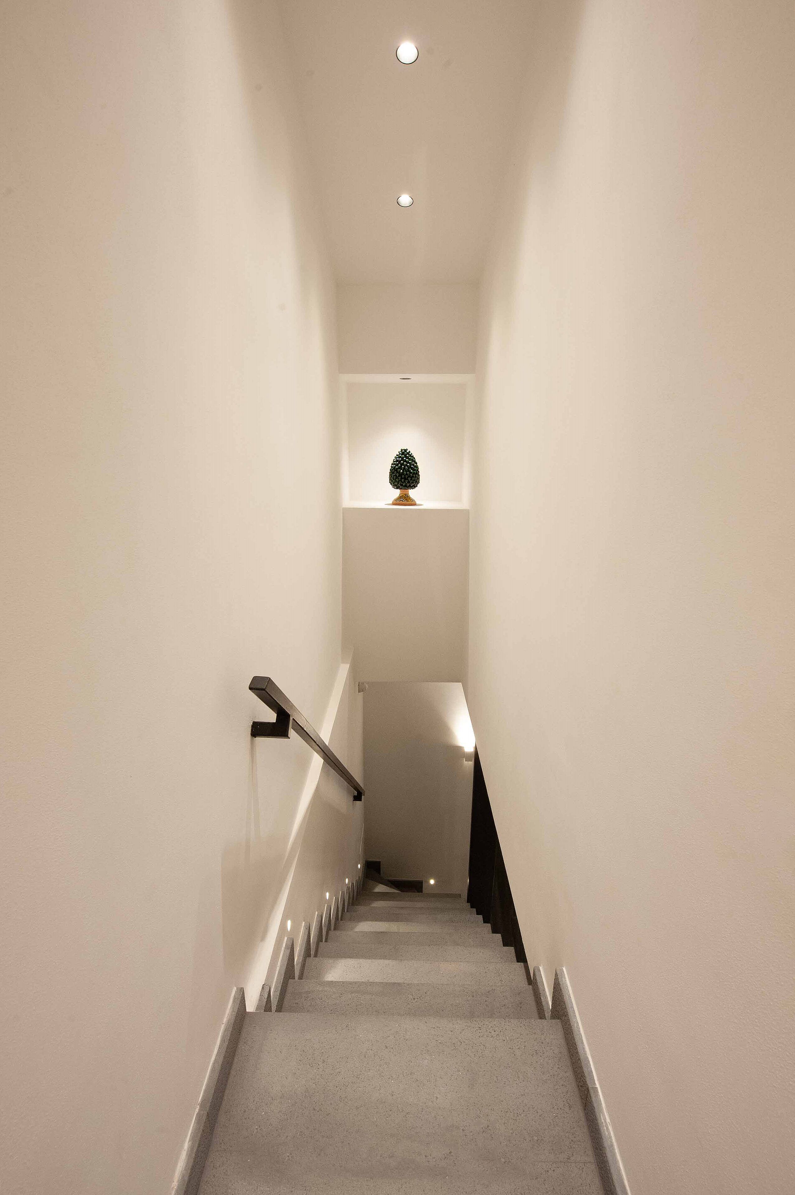 A narrow, well-lit staircase with a handrail on one side and a wall-mounted shelf with a small decorative item at the top.