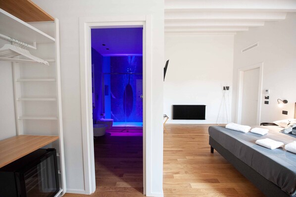 A modern hotel room with a wooden floor, a bed with white linens, a built-in wardrobe, and a bathroom with a blue-lit shower.