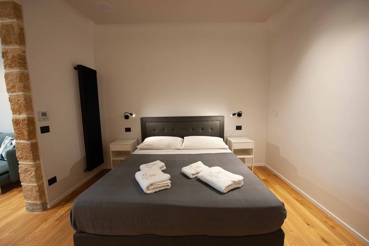 Een keurig opgemaakt bed met een donker hoofdbord, twee nachtkastjes met lampen, en een bakstenen muur aan de zijkant.