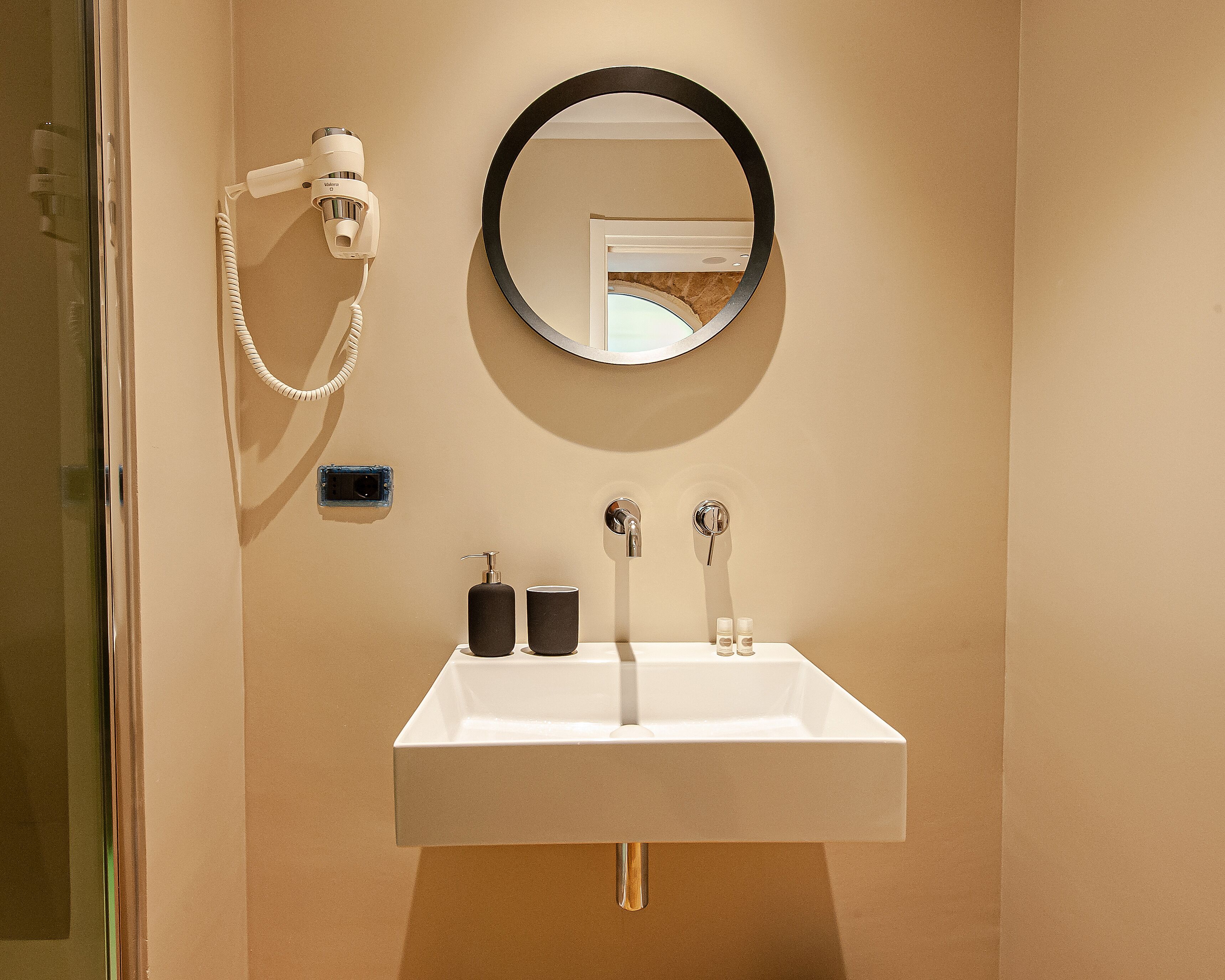 Un bagno con un lavandino bianco, uno specchio rotondo e un asciugacapelli fissato alla parete.