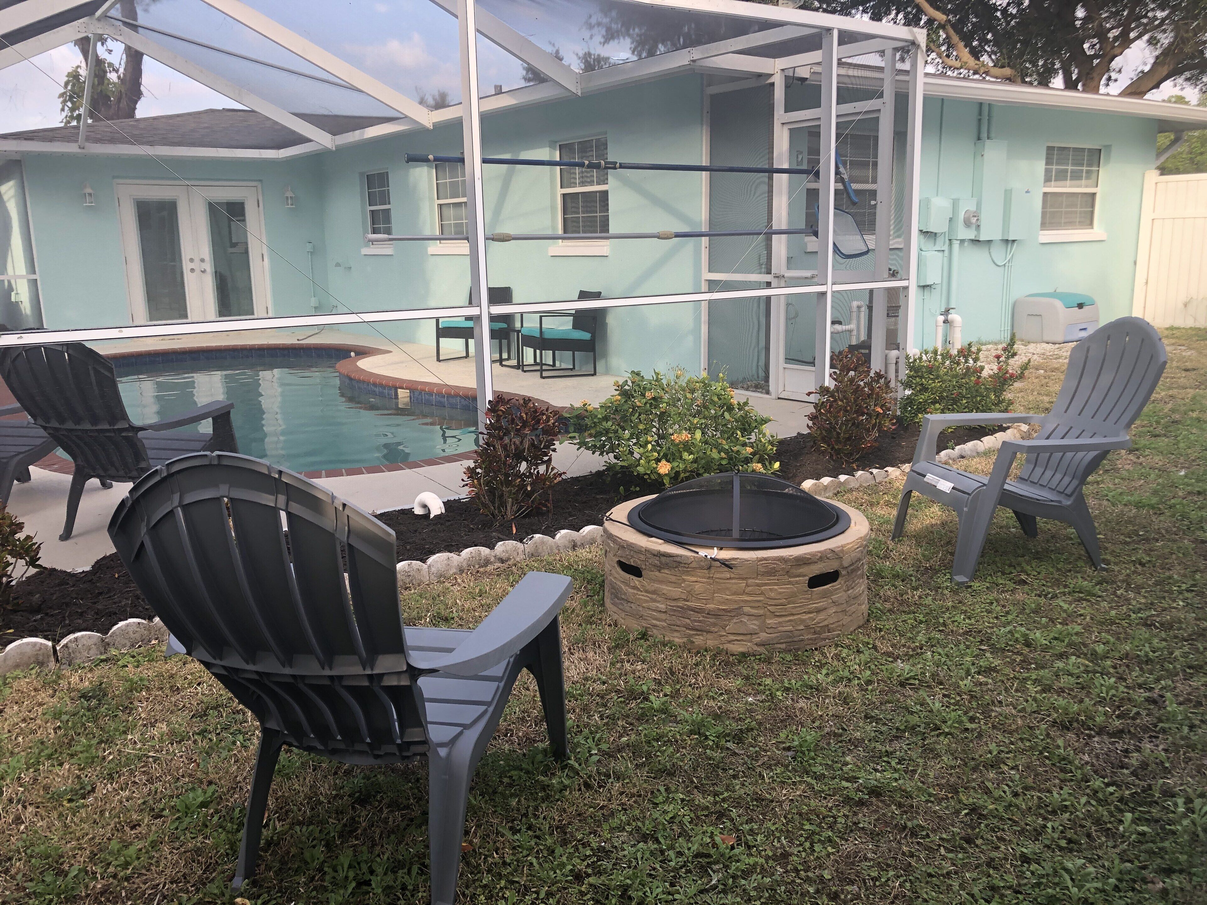 Un patio trasero con piscina, dos sillas Adirondack, un hoyo de fuego y un área de patio cubierta.