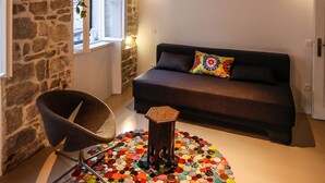 Un salon chaleureux avec un mur en pierre, un plafond en bois, un canapé noir agrémenté d’un coussin coloré, une table basse ronde ornée de pompons colorés et un fauteuil noir.