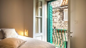 Une chambre à coucher avec un lit, des oreillers, une table de chevet, une fenêtre avec des rideaux, et une vue sur un mur en pierre et une rambarde verte à l’extérieur.