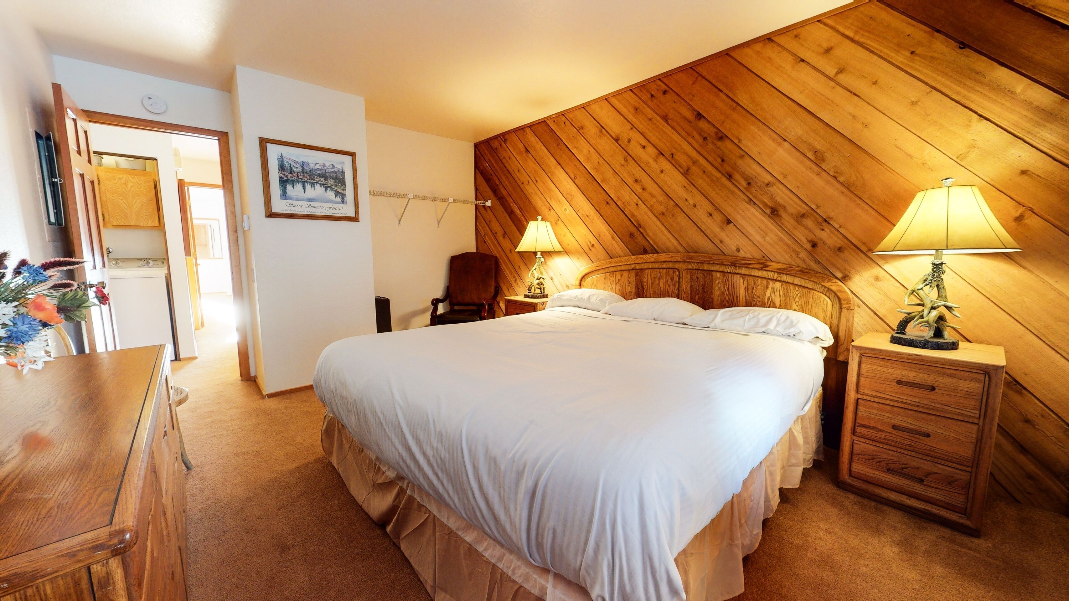 Un dormitorio con una cama grande, cabecera de madera, dos lámparas de noche, una mesita de noche, una silla, un aparador y un cuadro en la pared.