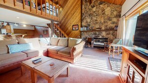 A cozy living room with a high ceiling, wooden beams, a stone fireplace, and a television. - Snowcreek III 448 Pet-Friendly, Spacious and Bright, Gorgeous Meadow Views by RedAwning (Mammoth Lakes)
