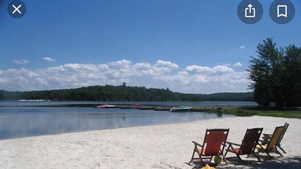 A beach with two red chairs and a yellow umbrella, a calm lake, and a forested island in the background. - Masthope  Community Charming Chalet 3 day min stay.  (Lackawaxen)